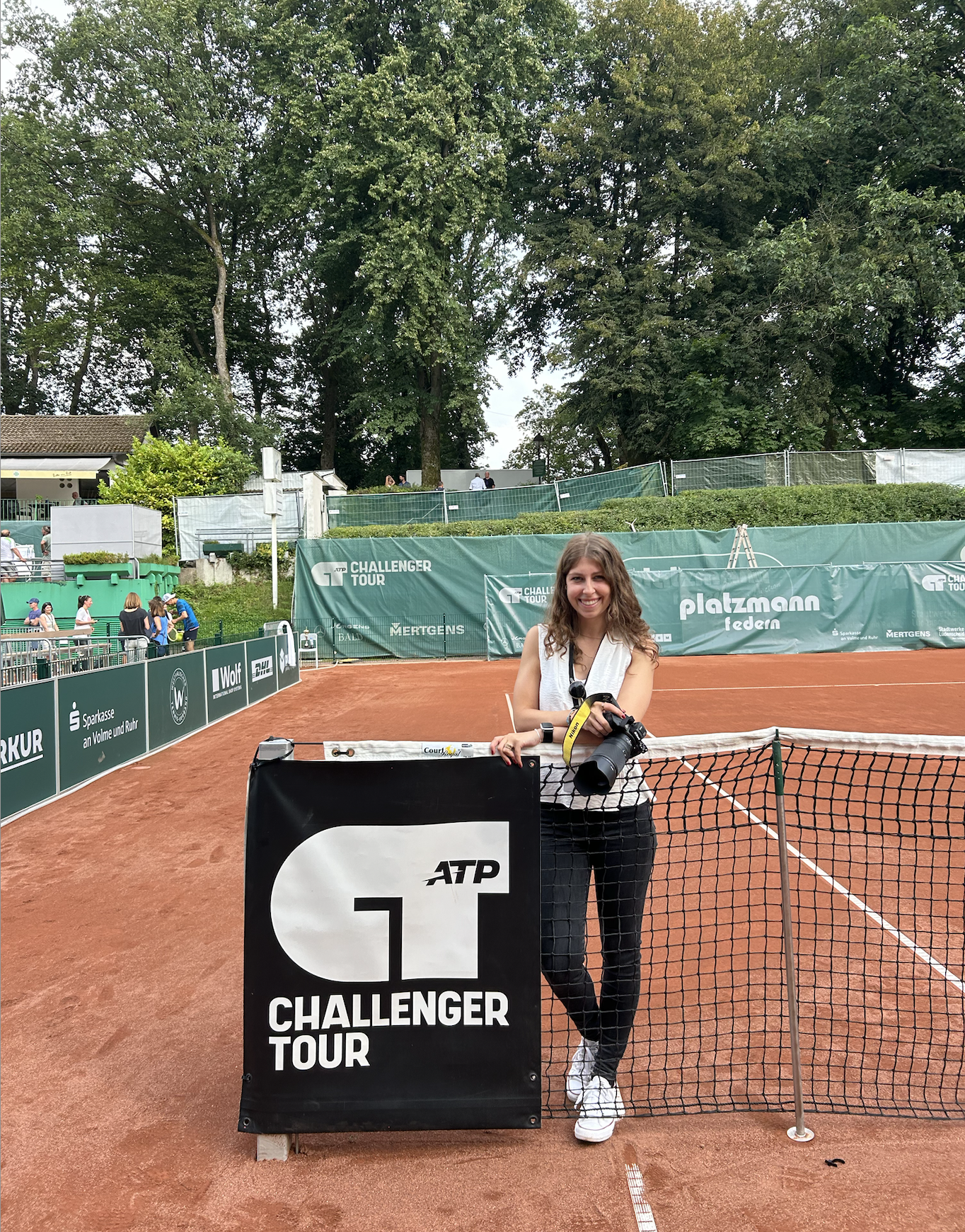 Sportfotografin steht draußen auf Ascheplatz vor Tennisnetz mit ATP Challenger Tour Schild und Kamera