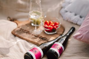 Weinflasche liegt mit Holzbrett, weinglaesern und glasschalen mit Erdbeeren und Weintrauben auf Picknickdecke in der sonne Produktaufmachung moods Brandkampagne