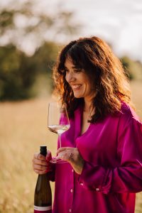 Geschaeftsportrait von Frau mit pinker bluse, die ihren wein "auf zack" von weinhaftig und Weinglas mit Weißwein als moods zum produktlaunch haelt