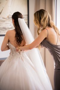 Brautjungfer mit Seidenkleid schließt Knoepfe des Brautkleides der Braut mit Schleier beim Getting Ready