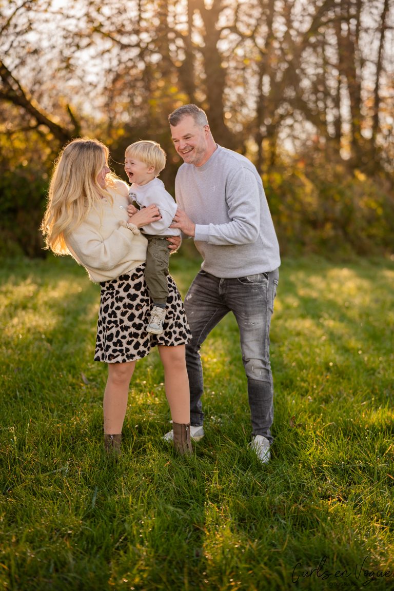 Familienfoto von Mama trägt lachenden Sohn auf dem Arm, der wird von Papa gekitzelt. Alle lachen auf Wiese mit warmer Sonne