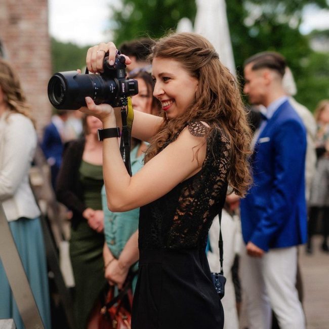 Hochzeitsfotografin mit schwarzem Hosenanzug haelt zwei Kameras und fotografiert Hochzeitspaar