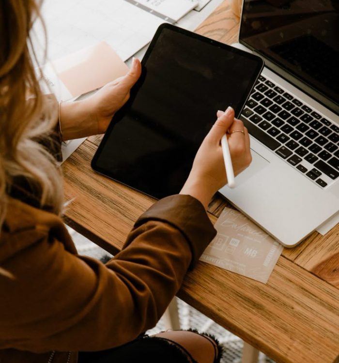 Frau ist social media Managerin und sitzt am Tisch und haelt tablet in der Hand. auf dem Tisch steht ein Laptop.