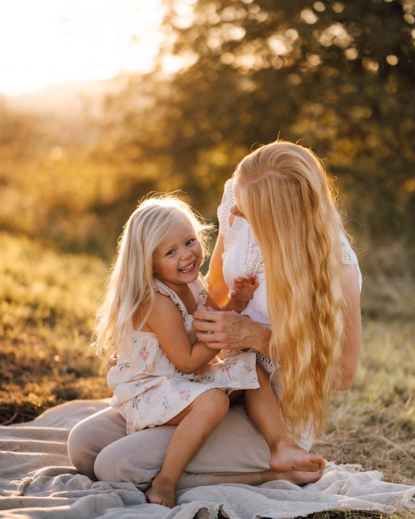 familienfoto von kleines maedchen sitzt auf schoß von mama und laechelt in kamera auf picknickdecke bei sonnenuntergang