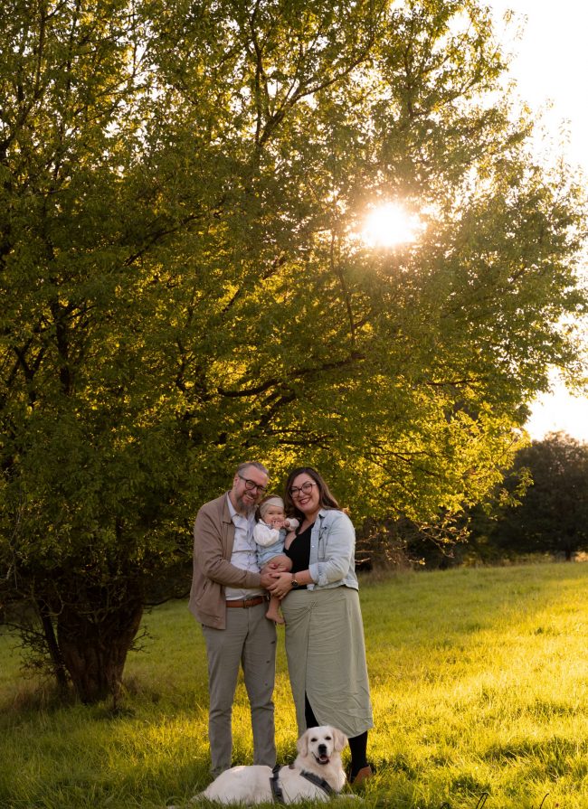 Familienportrait von Eltern mit Baby und Golden Retriever steht auf Wiese vor Baum wo Sonne durchscheint