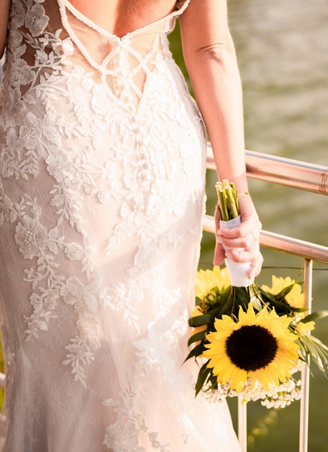 Braut traegt rückenfreies Brautkleid mit Blumenspitze und haelt Brautstrauß mit Sonnenblumen in der Hand