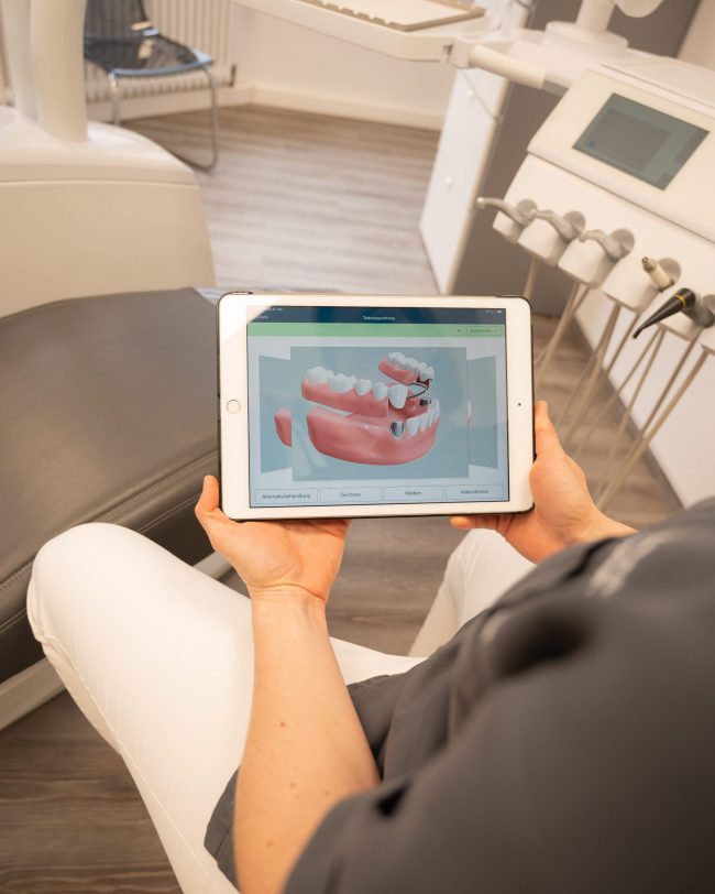 zahnaerztin sitzt im Behandlungsraum und haelt tablet in der hand welches neueste Kiefer und Zahntechnik zeigt