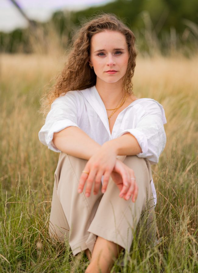 blonde Frau mit Naturlocken sitzt mit beiger Hose und weißer bluse auf wiese, harmonische und natuerliche farben